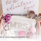 An open gift box for the bride to be showing a white cosmetic pouch with glittery 'MRS' lettering, makeup brushes, pink cosmetic sponges, pearl earrings and a floral accent headband. The inside lid reads 'let the adventure begin!' in rose-gold script.