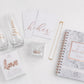Bridal subscription box flat lay arranged on a white background, including two glass champagne flutes with gold script reading Bride and Groom, two white vow books with gold script, a small clear ring box, a gold chain with a Mrs pendant, a rose gold LOVE balloon, and a wedding planning notebook